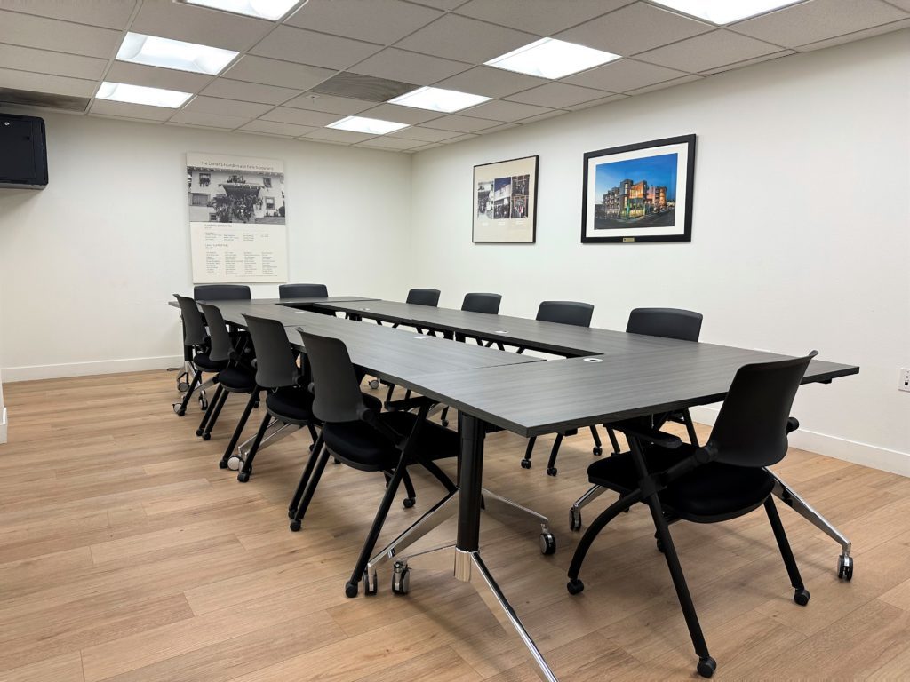 A room with white walls and a large table in the center of the room surrounded by dark grey chairs. There is 3 pieces of art on the walls.